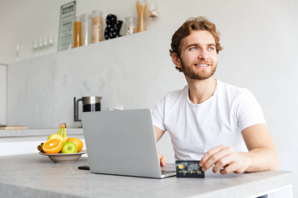Photo of a happy young bearded man at the table at home talking using laptop computer holding credit card. - Foto, Imagen