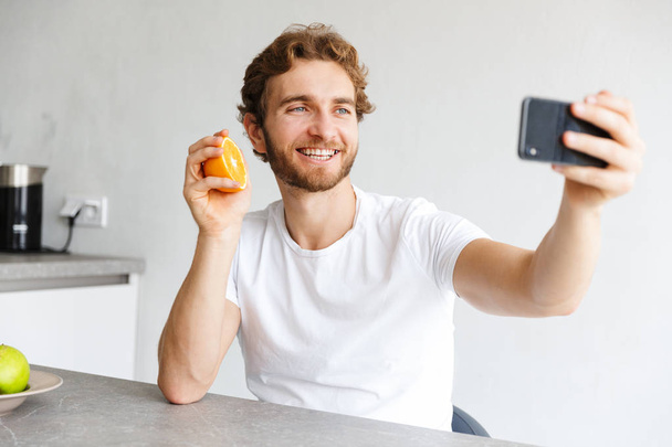 Photo of a happy young bearded man at the table at home make a selfie by phone with fruits. - Foto, imagen