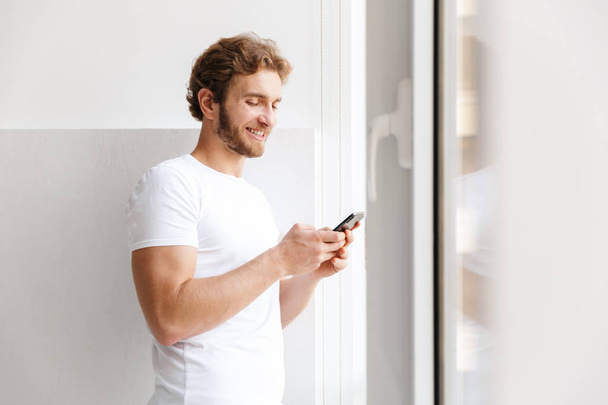 Smiling young man holding mobile phone, standing at the window at home - Photo, image