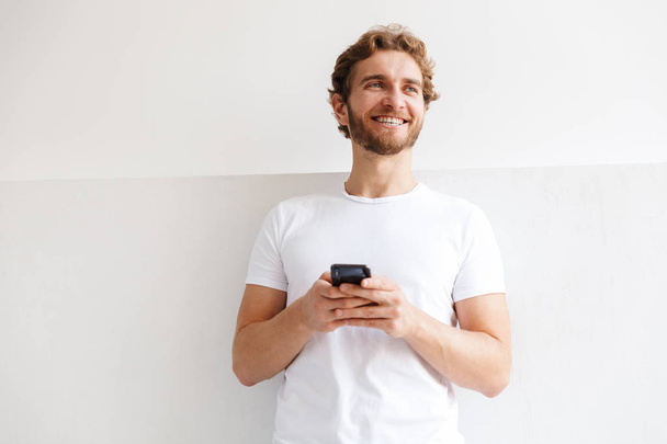 Smiling young man holding mobile phone, standing at the wall at home - Foto, immagini