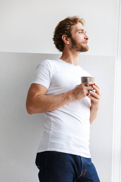 Smiling young man holding cup of tea, standing at the window at home - Foto, Imagem