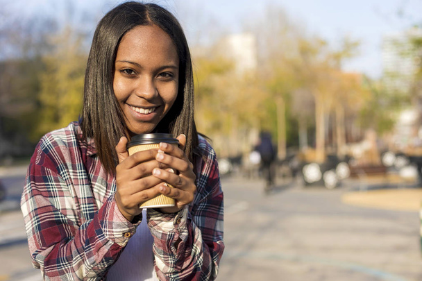Gülümseyen genç Afro-Amerikan kadın take away kahve tutarak ve güneşli bir günde kamera seyir sırasında caddenin ayakta - Fotoğraf, Görsel