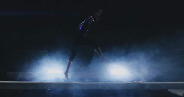 Chica gimnasta profesional realiza un salto con un giro en la viga de equilibrio en cámara lenta. Color azul humo en el gimnasio. Concursos de gimnasia
. - Imágenes, Vídeo