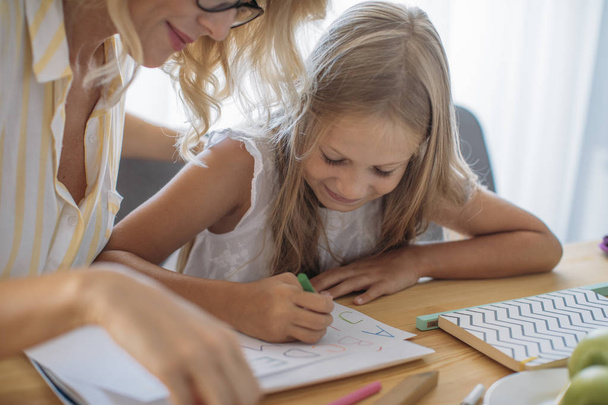 Cute Caucasian schoolgirl writing letters on paper with her mother helping. - Photo, image