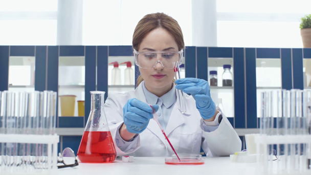Wissenschaftlerin in Schutzbrille steckt bei Experiment im Labor rote Flüssigkeit in Röhre - Filmmaterial, Video