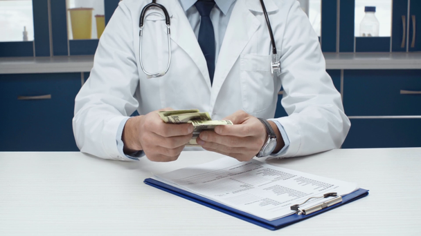 cropped view of male doctor in white coat sitting at table with diagnosis, counting and putting money in pocket - Footage, Video