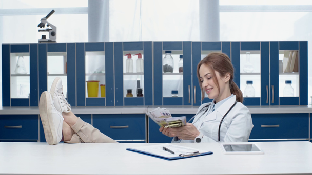 όμορφη χαμογελαστή γυναίκα γιατρό σε λευκό παλτό κάθεται στο τραπέζι και μετρώντας χρήματα στην κλινική - Πλάνα, βίντεο