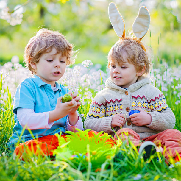 Cute adorable little kid boy making an egg hunt on Easter. Happy child searching and finding colorful eggs in domestic garden. Boy in spring clothes on cold day. Old christian and catholoc tradition. - Photo, Image