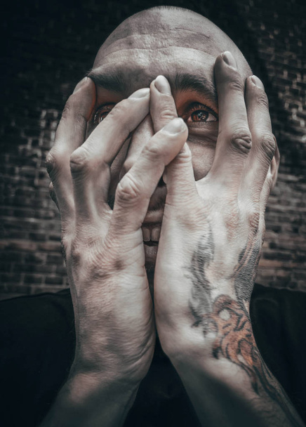 A close-up portrait of a bald man with a brutal appearance who covered his face with his tattooed hands - Foto, Imagen