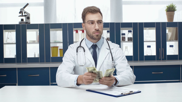 male doctor in white coat sitting at table with diagnosis, counting and putting money in pocket - Footage, Video