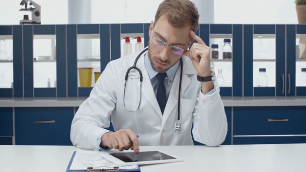guapo médico varón de capa blanca usando tableta digital y diagnóstico de escritura en la clínica
 - Metraje, vídeo