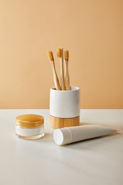 Bambuszahnbürsten im Halter, Zahnpasta in der Tube und Kosmetikcreme auf weißem Tisch und beigem Hintergrund - Foto, Bild