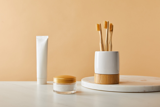 white holder with bamboo toothbrushes on marble round board, toothpaste in tube and cosmetic cream on table and beige background - Valokuva, kuva