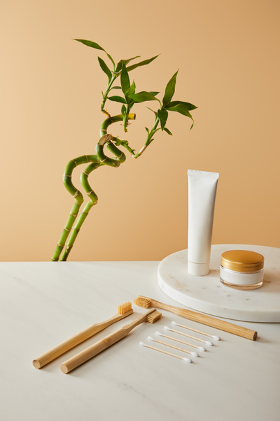 зубная паста в тюбике, косметические кремы, бамбуковые зубные щетки и палочки для ушей на столе, зеленый бамбук на бежевом фоне
 - Фото, изображение
