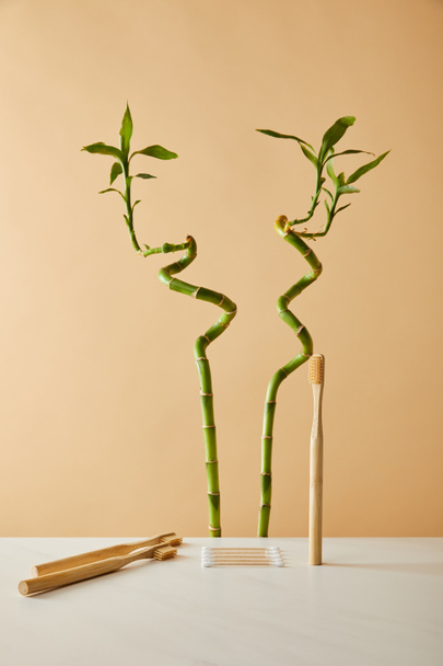 bamboe tandenborstel met oor stokken op witte tafel en groene bamboe op beige achtergrond - Foto, afbeelding
