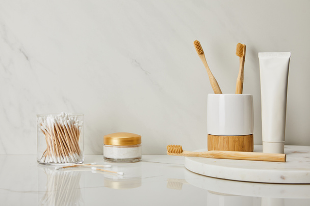 tandpasta in de buis, houder, bamboe tandenborstels, cosmetische crème en oor blijven hangen in glas op witte marmeren achtergrond - Foto, afbeelding