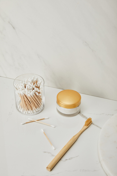ear sticks in glass, cosmetic cream and bamboo toothbrush on white marble background - 写真・画像
