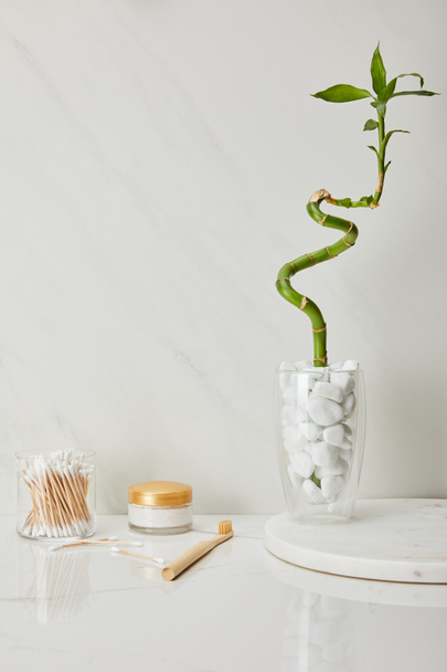 ear sticks in glass, cosmetic cream and toothbrush near bamboo stem in vase on white marble background - Zdjęcie, obraz