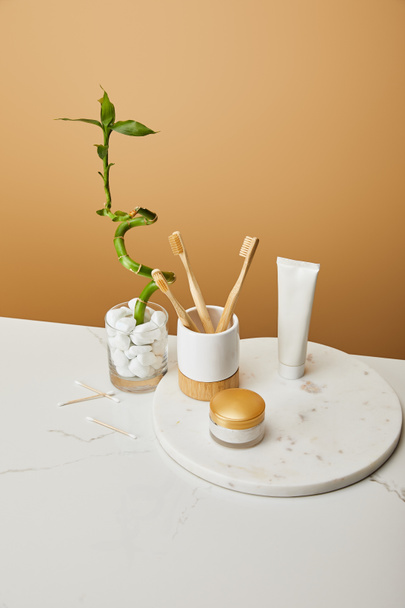 spazzolini di bambù in supporto, crema cosmetica e dentifricio in tubo su tavola rotonda vicino al gambo di bambù verde in vaso su fondo beige
 - Foto, immagini