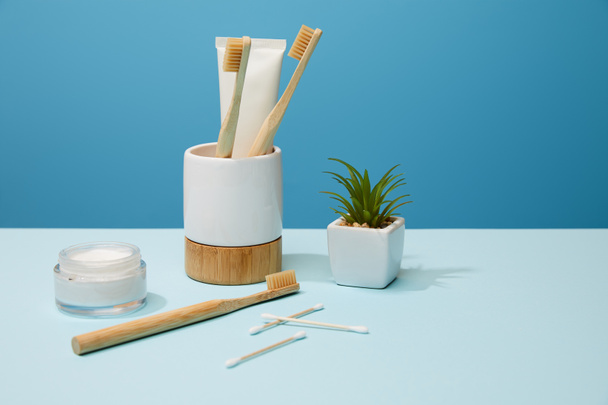 Halter für Zahnbürsten, Kosmetikcreme und Pflanze im Topf auf Tisch und blauem Hintergrund - Foto, Bild