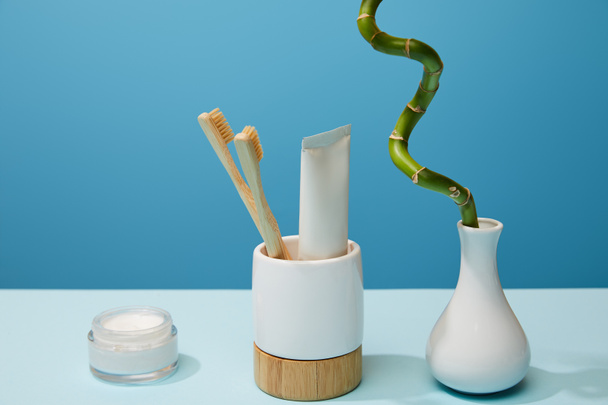 soporte para cepillos de dientes, crema cosmética y tallo de bambú en jarrón sobre mesa y fondo azul
 - Foto, Imagen