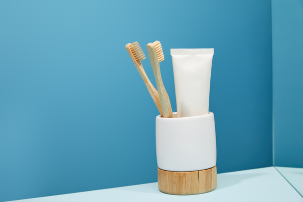 soporte con cepillos de dientes de bambú, pasta de dientes en tubo y espejo sobre mesa y fondo azul
 - Foto, Imagen