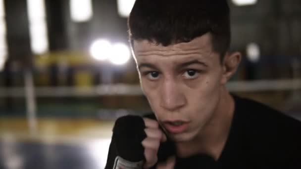 Young man in black T shirt fulfills blows in black boxing bandages and looking seriously at camera in the gym. Close up tired from workout boxer. Boxing ring, soffits and gym on the background. Close - Filmagem, Vídeo