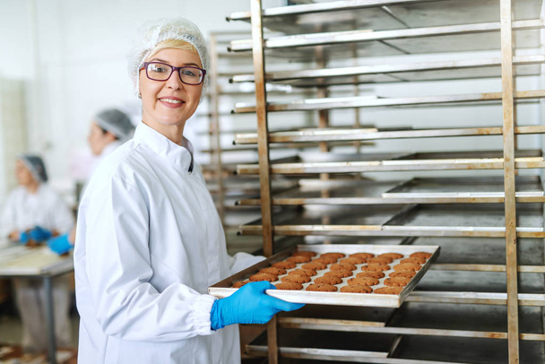 Усміхнена блондинка-жінка-працівник з окулярами і в стерильній формі кладе лоток з смачним печивом в стійку
. - Фото, зображення