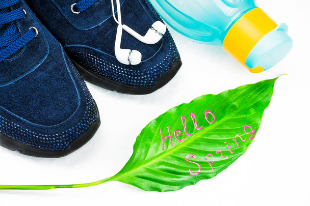 Doing fitness in the spring, sports concept. Green leaf with signature Hello Spring. Headset to listen to music. Blue, women's sneakers and sport gloves for the simulator. Eco bottle in bright colors for water. The background is light, large pane. - Photo, Image