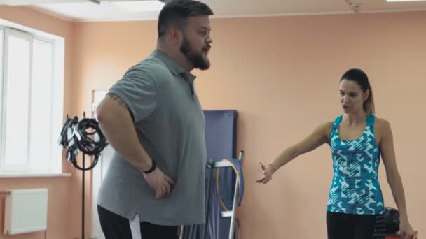 Personal training from athletic girl for fatboy with big abdomen in sportswear. Fat man training with woman instructor and doing exercises in fitness center. Thick obese guy together with personal - Πλάνα, βίντεο