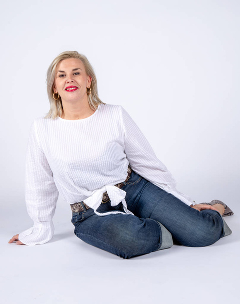 Séance studio photographique pour une femme de 50 ans assise sur le sol et vêtue de jeans et chemise blanche
. - Photo, image
