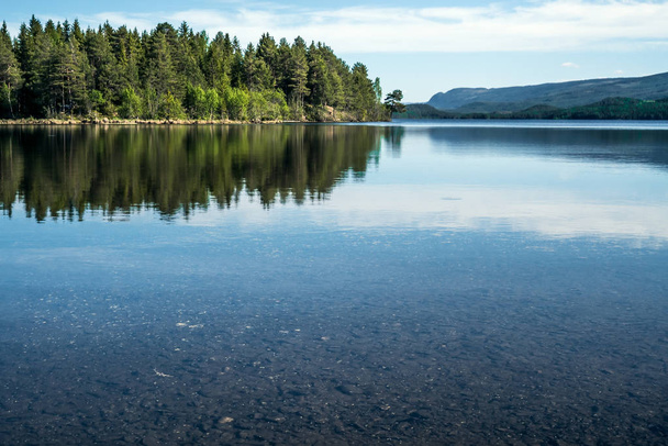 ein ruhiger Sommermorgen mit Blick auf den kristallklaren, ruhigen See mit Kieselsteinen im Boden und grünem Wald, der sich auf der Wasseroberfläche mit Bergen am Horizont spiegelt - Konzept der stillen Meditation und Erholung in der Natur. - Foto, Bild