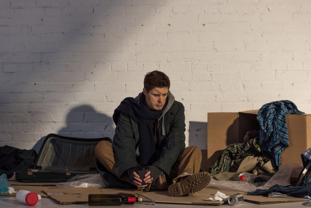 Depressiver Obdachloser sitzt auf Pappe und hält Pappbecher in der Hand - Foto, Bild