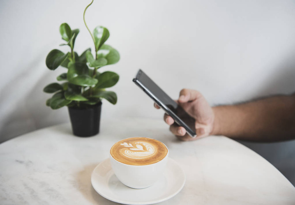 Hand mit Smartphone in den sozialen Medien mit Kaffee-Latte-Tasse im Café, Lebensstil im Alltag.  - Foto, Bild