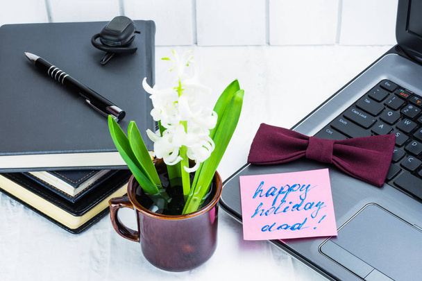 Business concept in the office. Flower arrangement for congratulating dad on the holiday. Work environment, happy dad. On a light background - a laptop, several business notebooks, a black pen. Headset for phone, bow tie for burgundy business suit. C - Photo, Image