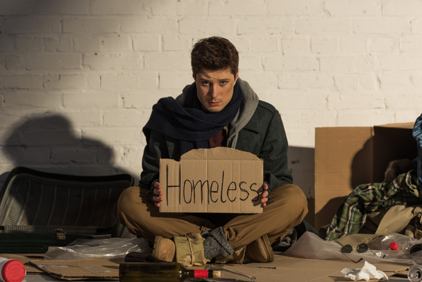 sad man sitting on rubbish dump and holding cardboard card with "homeless" handwritten text - Photo, Image