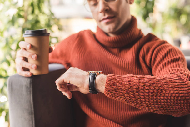 selective focus of pensive man holding paper cup while looking at watch in cafe - Photo, Image