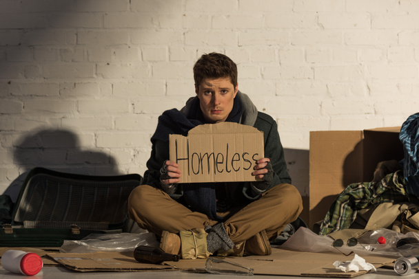 Obdachloser Mann hält Pappkarte mit "obdachlos" handgeschriebenem Text in der Hand, während er auf Müllkippe sitzt - Foto, Bild
