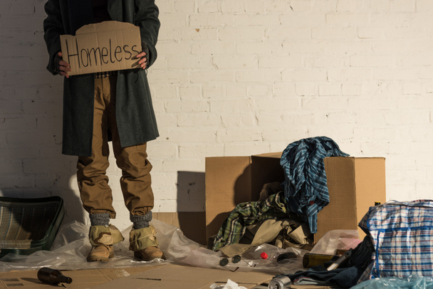 vista parcial del hombre sin hogar parado en el basurero y sosteniendo un pedazo de cartón con inscripción manuscrita "sin hogar"
  - Foto, Imagen