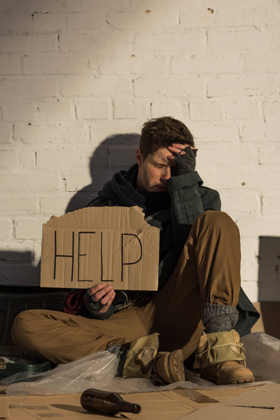 Verzweifelter Obdachloser sitzt an Ziegelmauer und hält Stück Pappe mit "Hilfe" -Aufschrift in der Hand - Foto, Bild
