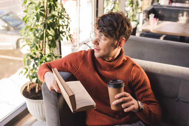 pensive young man in glasses holding book and paper cup in hands - Photo, image