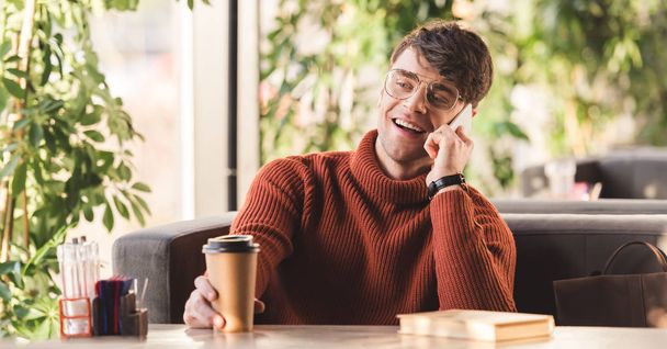 selective focus of cheerful man in glasses talking on smartphone near paper cup with coffee and book  - 写真・画像