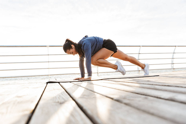 Side view of a concentrated sportswoman doing plank exercises on a fitness mat at the beach - Fotó, kép