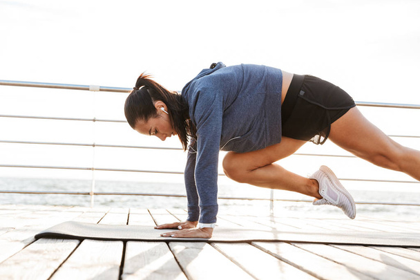 Side view of a concentrated sportswoman doing plank exercises on a fitness mat at the beach - Foto, afbeelding