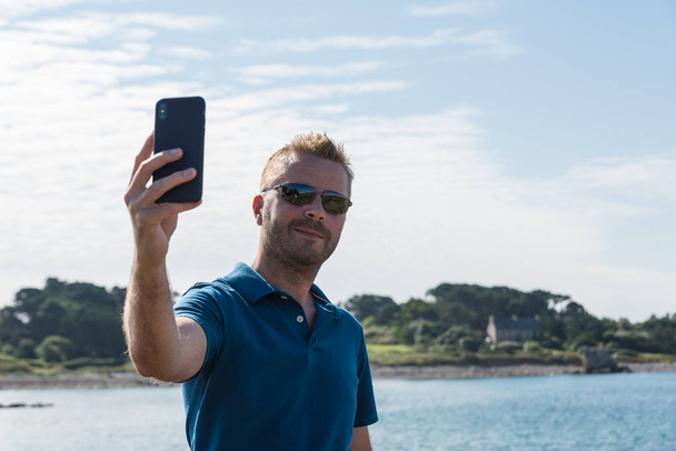 Мужчина делает селфи со смартфоном на берегу озера
 - Фото, изображение