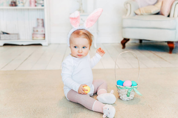 Pikkutyttö pupunkorvissa pääsiäispäivänä. Tyttö, jolla on kori, jossa on maalattuja munia, istuu lattialla kotona, hauskaa pääsiäismunan metsästyksessä. Hyvää pääsiäistä loma juhla kevät konsepti
 - Valokuva, kuva