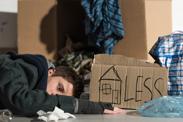 Depressiver Obdachloser liegt auf Pappe in Müllhalde, mit Haussymbol und "weniger" Aufschrift auf Pappkarte - Foto, Bild