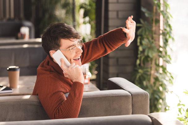 joven alegre en gafas hablando en el teléfono inteligente mientras agita la mano en la cafetería
 - Foto, imagen