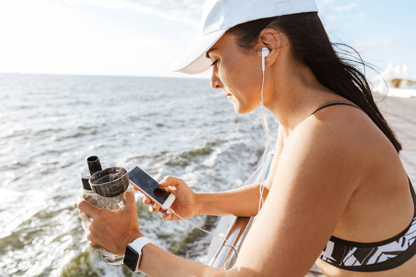Image of a beautiful young sports fitness woman using mobile phone drinking water at the beach outdoors listening music with earphones. - Photo, Image