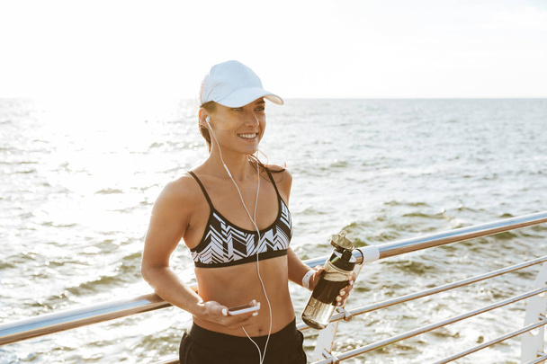 Image of a beautiful young sports fitness woman using mobile phone drinking water at the beach outdoors listening music with earphones. - Фото, изображение
